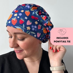 May include: A blue scrub hat with a colorful strawberry, cherry, and blueberry print. The hat has a gathered elastic band and includes a ponytail tie.