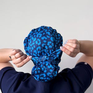 May include: A blue scrub cap with a leopard print pattern in shades of blue. The cap is tied at the back. The person in the image is wearing a navy blue scrub top.