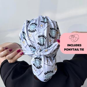 May include: A light blue surgical scrub cap with a repeating pattern of surgical masks, daisies, and the words "General Surgery." The cap includes a ponytail tie. The cap is tied in a knot at the back.