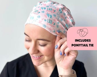 May include: Pink and blue patterned scrub hat with the text "L&D nurse" repeated throughout. The hat is worn by a person with blue nail polish. The image includes a pink box with the text "Includes Ponytail Tie".