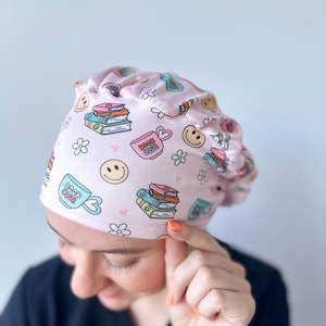 May include: Pink surgical scrub hat with a book club theme. The hat features a pattern of books, coffee cups, and smiley faces. The text "Book Club" is printed on the hat.