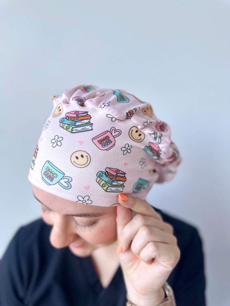 May include: Pink surgical scrub hat with a book club theme. The hat features a pattern of books, coffee cups, and smiley faces. The text "Book Club" is printed on the hat.