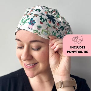May include: A white scrub cap with a colorful pattern of potted plants, including ferns and succulents. The cap is shown with a ponytail tie. The cap is worn by a person wearing a black top.