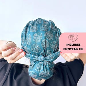 Op de afbeelding: Teal stoffen hoofdbedekking met een wit bladerpatroon, vastgeknoopt. De afbeelding bevat een roze rechthoek met de tekst "INCLUDES PONYTAIL TIE" en een tekening van een paardenstaartstrik.