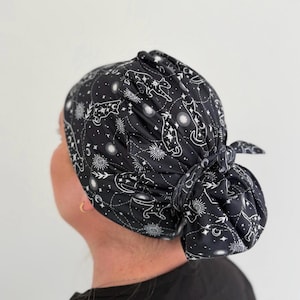 以下が含まれることがあります： 星、太陽、月、そして猫のシルエットが描かれた白い天体模様の黒い手術用キャップ。生地は後ろでギャザーが寄せられ、結び目で固定されており、髪を覆うためのボリュームのある形状を作り出しています。
