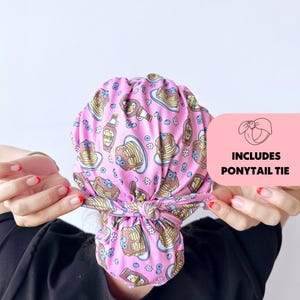 Op de afbeelding: Een roze scrubmuts met een patroon van pannenkoeken, bosbessen en siroopflesjes. De muts bevat een paardenstaartband. De tekst "INCLUDES PONYTAIL TIE" staat op een roze achtergrond.