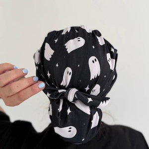 May include: A black scrub cap featuring a repeating pattern of white ghosts and small four-pointed stars. The cap is gathered at the back and secured with a tied fabric knot, creating a rounded shape that covers the head and hair completely.