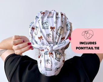 May include: A white scrub cap with a pattern of books, bows, and flowers. The cap is tied at the back and includes a ponytail tie. The text "INCLUDES PONYTAIL TIE" is in a pink box.