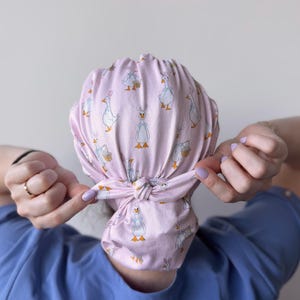 Op de afbeelding: Een roze scrubcap met een herhaald patroon van cartoon ganzen met strikjes en manden. De cap is achteraan geknoopt. De achtergrond is lichtgrijs.