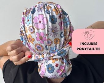 May include: A white floral print scrub cap with pink, blue, and orange flowers. The cap is tied in a knot at the back. The image includes the text "INCLUDES PONYTAIL TIE".