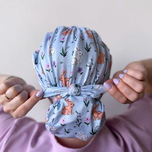 Op de afbeelding: Een lichtblauwe scrubcap met een patroon van oranje konijnen, witte konijnen, paarse bloemen en groene bladeren. De cap is aan de achterkant vastgeknoopt.