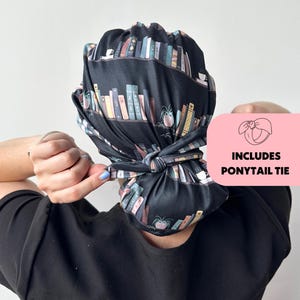 Op de afbeelding: Een zwarte scrubcap met een herhaald patroon van kleurrijke boeken, koffiekopjes en potplanten. De cap is achterin geknoopt. Een roze vierkant in de afbeelding leest "INCLUDES PONYTAIL TIE".