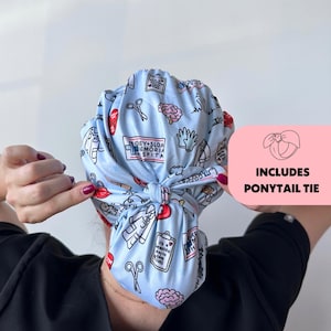 May include: A light blue scrub cap with a repeating pattern of medical-themed illustrations, including scissors, brains, and medical charts. The cap includes a ponytail tie. The text "Gey+Sloa Memorial Spita" is visible on the cap.