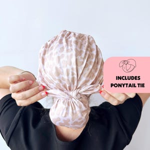 Op de afbeelding: Een lichtroze en beige patroon hoofddoek vastgebonden in een knot. De hoofddoek wordt vastgehouden met de handen van een persoon die een zwarte top draagt. De afbeelding bevat een roze vierkant met de tekst "INCLUDES PONYTAIL TIE".
