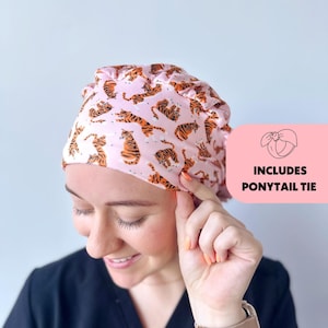May include: Pink surgical scrub hat with a tiger print pattern. The hat has a ponytail tie.