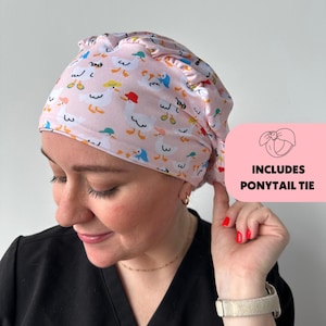 May include: A pink scrub cap with a pattern of cartoon ducks wearing hats. The cap has a ruffled edge and includes a ponytail tie. The text "INCLUDES PONYTAIL TIE" is in a pink box.