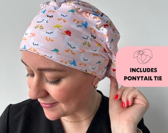 May include: A pink scrub cap with a pattern of cartoon ducks wearing hats. The cap has a ruffled edge and includes a ponytail tie. The text "INCLUDES PONYTAIL TIE" is in a pink box.