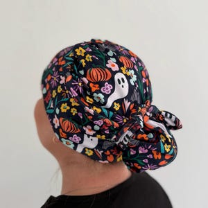 May include: A dark navy blue scrub cap featuring a repeating pattern of white ghosts, orange pumpkins, and colorful flowers in shades of pink, yellow, and teal. The cap has a gathered back with a tied knot detail to secure the fabric in place.