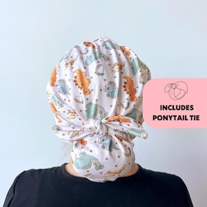 May include: A white fabric head covering with a dinosaur pattern in orange, blue, and green. The head covering includes a ponytail tie. The design features various dinosaur illustrations and small star accents.