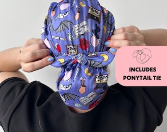 May include: A blue surgical scrub cap with a repeating pattern of bats, books, roses, and moons. The cap includes a ponytail tie. The cap is being held up by a person.