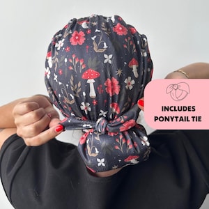 Op de afbeelding: Een donkergrijze stoffen hoofdbedekking met een bloemen- en paddenstoelenpatroon. Het ontwerp bevat rode bloemen, witte bloemen en witte paddenstoelen. De tekst "INCLUDES PONYTAIL TIE" staat op een roze achtergrond.