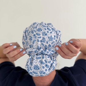 May include: A white scrub cap featuring a repeating blue line-art pattern of small ghosts holding bouquets of flowers. The cap has a gathered back design with a fabric tie closure knotted in the center, securing the fabric in a bun-style shape.