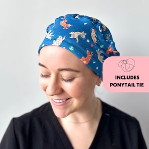 May include: A blue scrub cap with a pattern of cartoon cats in space. The cap has a ruffle around the edge and includes a ponytail tie.