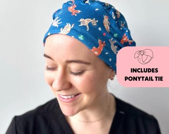 May include: A blue scrub cap with a pattern of cartoon cats in space. The cap has a ruffle around the edge and includes a ponytail tie.