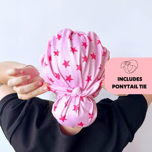 Op de afbeelding: Roze hoofdwikkel met een herhalend patroon van rode sterren. De hoofdwikkel is aan de achterkant in een knoop geknoopt. De afbeelding bevat een roze vak met de tekst "INCLUDES PONYTAIL TIE".