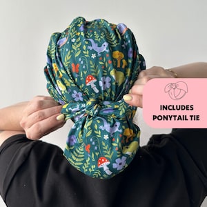 May include: A green fabric hair wrap with a whimsical pattern of mushrooms, flowers, butterflies, and lizards. The wrap is tied in a knot at the back. The image includes a pink box with the text "INCLUDES PONYTAIL TIE".
