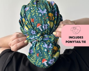 May include: A green fabric hair wrap with a whimsical pattern of mushrooms, flowers, butterflies, and lizards. The wrap is tied in a knot at the back. The image includes a pink box with the text "INCLUDES PONYTAIL TIE".