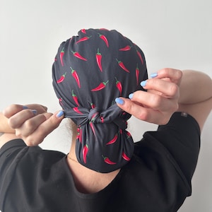 Puede incluir: Un gorro negro con un estampado de chiles rojos. El gorro está atado en la parte posterior. La persona lleva una camisa negra.