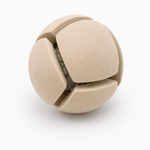 May include: A spherical object composed of several light beige, curved segments with a visible concentric circular texture. The segments are arranged to form a ball shape, revealing a white internal structure through the gaps between the pieces.