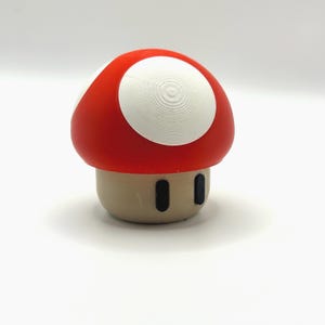 May include: A mushroom-shaped figure with a rounded red cap featuring white circular spots. The base is a light tan colour and features two small, vertical black oval shapes on the front, resembling eyes or decorative markings.