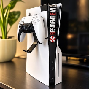 May include: A white and black games console with a matching wireless controller mounted on a black stand. The console features a vertical black panel with the text Resident Evil in white and red letters, along with a red and white umbrella logo emblem.