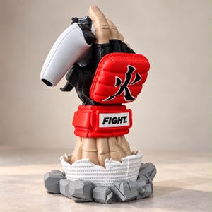 May include: A sculpted figurine of a hand wearing a black and red martial arts glove with a Japanese character, holding a white game controller. The wrist features a red band with the text FIGHT. The base is white and surrounded by grey rocks.