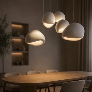 May include: A cluster of five off-white, organic, pebble-shaped pendant lights suspended at varying heights. Each fixture features a smooth, matte surface with a wide, downward-facing opening that emits a warm, soft glow from an internal light source.