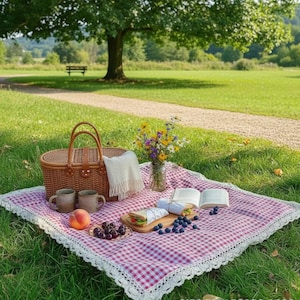 Può includere: Un cestino da picnic in vimini marrone intrecciato con cinghie in pelle, un panno bianco con frange, un vaso di vetro con fiori di campo, due tazze in ceramica, una pesca, una ciotola di ciliegie, due panini e un libro aperto su una tovaglia a quadri.