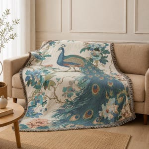 Puede incluir: Una manta tejida que presenta un gran pavo real con plumaje azul y verde posado en una rama, rodeado de motivos florales en tonos azul, verde y melocotón sobre un fondo crema, acabado con un borde de flecos en todos los lados.