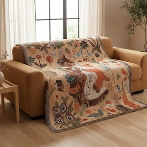 Peut inclure: Un plaid tissé sur fond beige orné d'un motif détaillé représentant un chat endormi, un renard, diverses fleurs colorées, des champignons, des papillons et de petits oiseaux. La couverture présente des bords à franges et recouvre un canapé beige.
