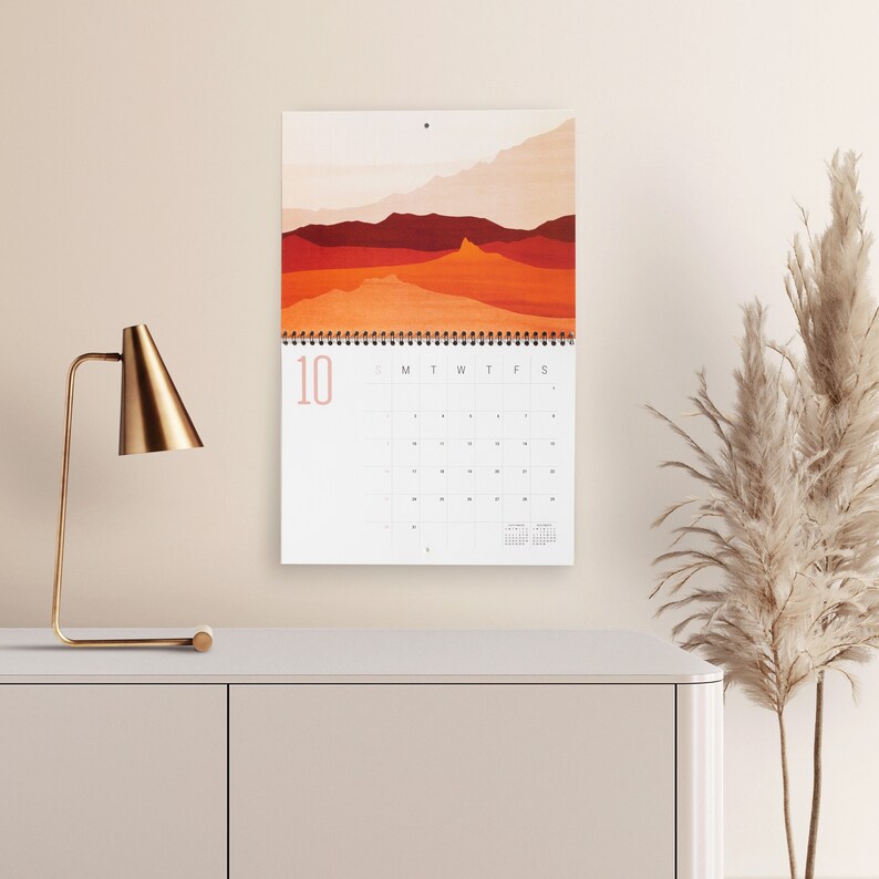 Wall calendar for 2024 with bright colorful abstract landscapes. Coil bound with pre-drilled hole for easy hanging. Calendar grid with space for notes. Opens up to 11x17 inches size. Month of October shown here with calendar hanging over a sideboard.