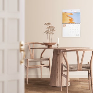Wall calendar for 2024 with bright colorful abstract landscapes. Coil bound with pre-drilled hole for easy hanging. Calendar grid with space for notes. Opens up to 11x17 inches size. Month of July shown here with calendar hanging in the dinning room.