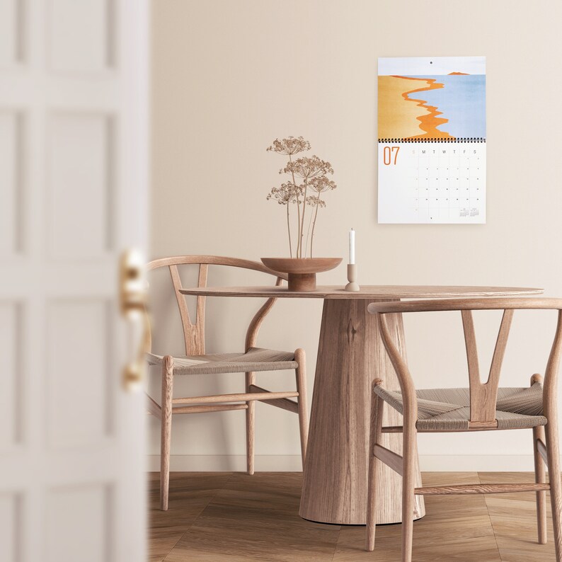 Wall calendar for 2024 with bright colorful abstract landscapes. Coil bound with pre-drilled hole for easy hanging. Calendar grid with space for notes. Opens up to 11x17 inches size. Month of July shown here with calendar hanging in the dinning room.