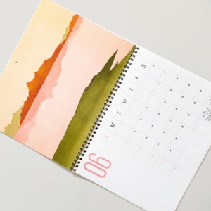 Wall calendar for 2024 with bright colorful abstract landscapes. Coil bound with pre-drilled hole for easy hanging. Calendar grid with space for notes. Opens up to 11x17 inches size. Month of June shown here.