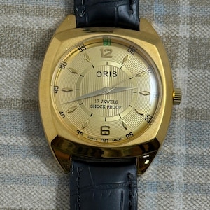 Puede incluir: Un reloj de pulsera Oris en tono dorado con esfera dorada texturizada, que incluye el texto ORIS, 17 JEWELS, SHOCK PROOF y SWISS MADE. El reloj tiene una caja dorada, marcadores de hora dorados y una correa de cuero negro con relieve.