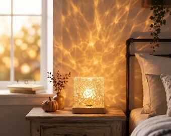 May include: A square glass lamp with a textured, rippled surface sits on a wooden base, emitting a warm, golden light that casts intricate, water-like patterns onto the wall. The lamp is illuminated, creating a soft glow in the room.