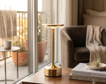 May include: A tall, slender table lamp with a brushed gold finish, featuring a circular base, a thin central stem, and a flat, disc-shaped top that emits a soft, downward-facing light from its underside, standing on a wooden surface.