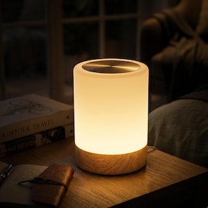 Puede incluir: Una lámpara de mesa cilíndrica con una pantalla de vidrio blanco esmerilado que emite un brillo cálido, rematada con una tapa circular de metal plateado y apoyada sobre una base de madera clara con vetas visibles y diseño limpio.