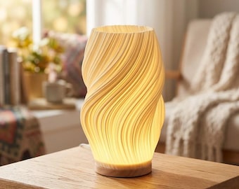 May include: A cylindrical table lamp with a cream-colored, 3D-printed shade featuring a vertical, wavy, ribbed texture. The lamp emits a warm, soft glow from within and is mounted on a circular, light-toned wooden base that sits flat.