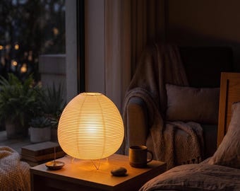 May include: A round, off-white paper lantern lamp with horizontal ribbing, supported by thin metal legs. The lamp emits a warm, soft glow and sits on a wooden bedside table next to a dark ceramic mug and a small, smooth, dark stone object.